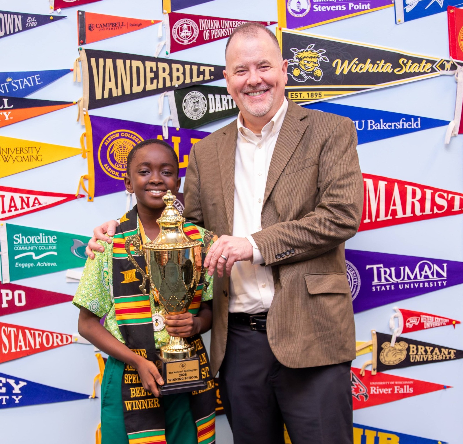 Eugene Mensah Osei poses with his championship trophy alongside an adult companion, in front of a wall of college pennants.
