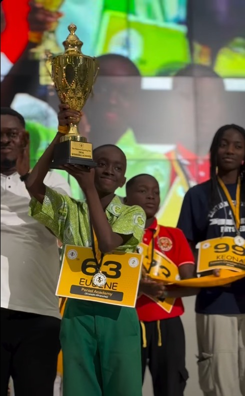 Eugene shakes hands with an event official on the championship stage while holding his trophy, with seated attendees behind.